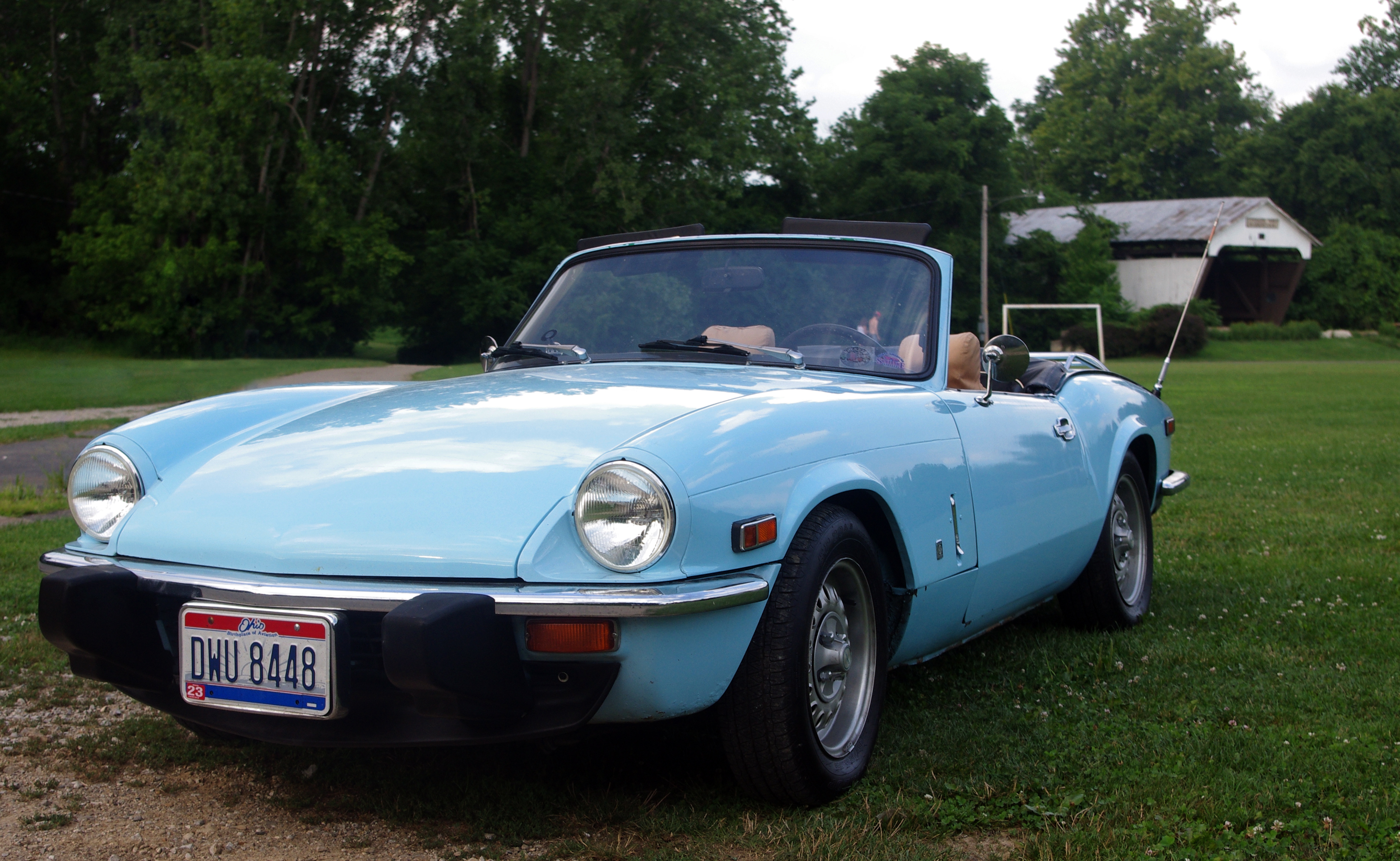 Light blue Triumph Spitfire convertible with its top down, photographed on grass in front of trees